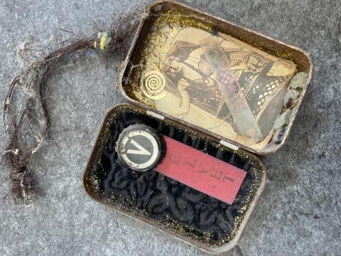 A small square tin sits on a grey countertop. There is black material on one side of the tin, on top of which is a larger black bottlecap with a capital "V" on it. Below the V is a piece of red cardstock with letters; together with the bottlecap, it spells out "Velvet". The other side is a sepia-toned image of a woman.