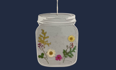 d lA suncatcher shaped like a mason jar with pressed flowers inside, against a blue backdrop.